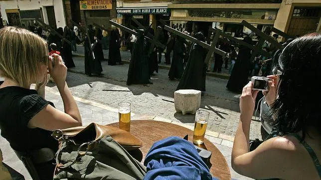 Dos turistas toman un refrigerio en un bar mientras toman fotografias de una procesión sevillana