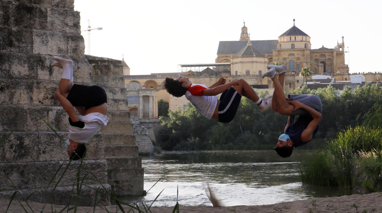 Unos jóvenes con mascarillas practican deporte bajo el puente romano de Córdoba con la mezquita catedral al fondo