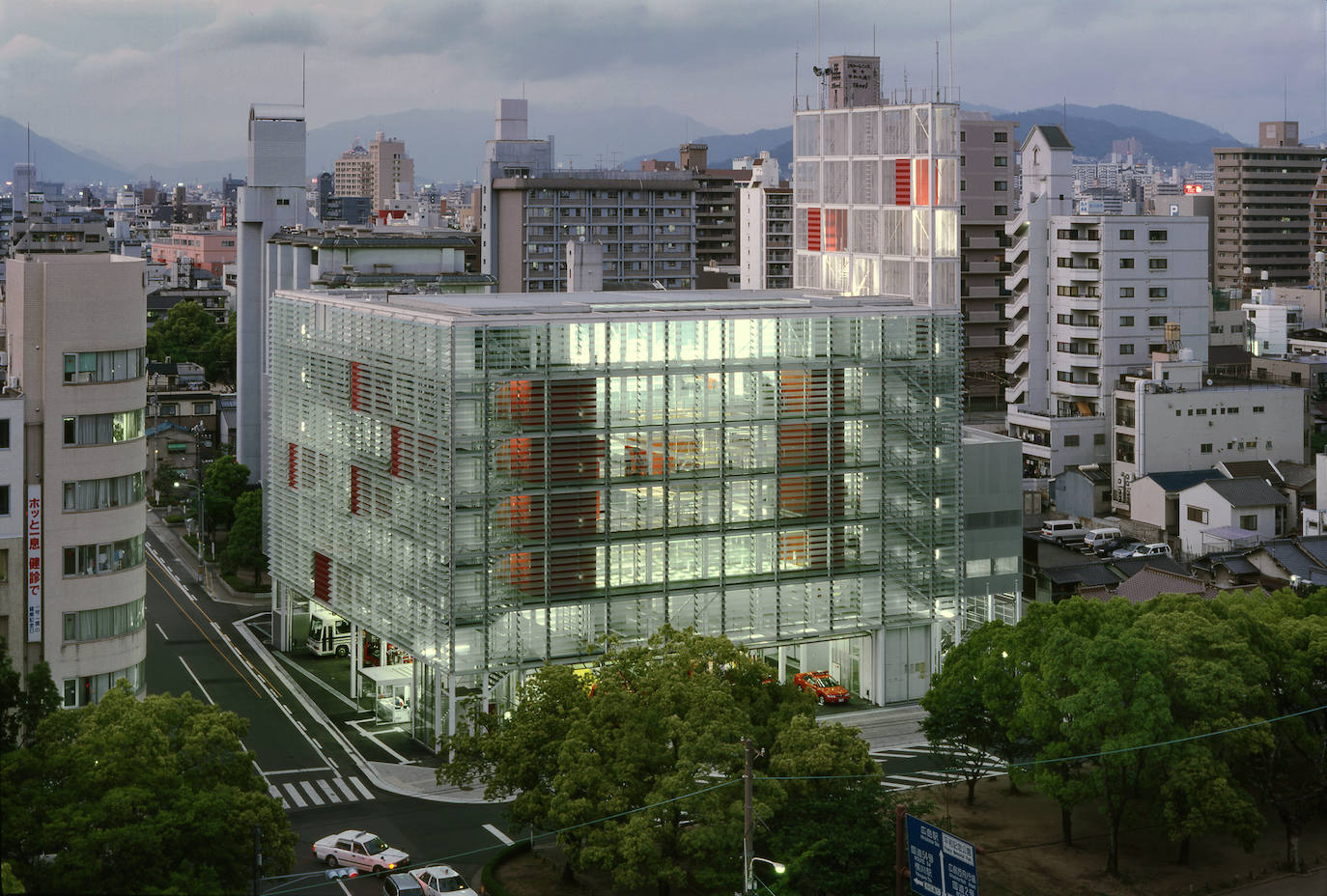 Estación de bomberos Hiroshima Nishi, Hiroshima (Japón), 2000. Fachada, paredes interiores y suelo están construidos en cristal, creando un gran volumen transparente que permite a todos los transeúntes ver los entrenamientos y actividades que los bomberos desempeñan en el interior, expresando el nexo de conexión entre este cuerpo de funcionarios y ciudadanos. «Un cuartel de bomberos debería jugar un importante papel en la formación de una comunidad local», señala Yamamoto. El vestíbulo y una terraza en la cuarta planta están abiertas al acceso público. Un atrio central funciona como núcleo desde el que se articulan todo el resto de instalaciones. La casi totalidad del edificio consiste en espacios separados mediante paredes de cristal.