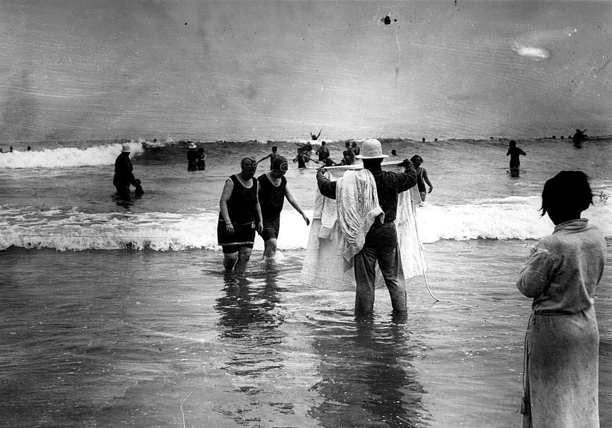 Un bañero espera a dos señoras mientras otros bañan a niños y mayores junto a las olas en San Sebastián en 1918