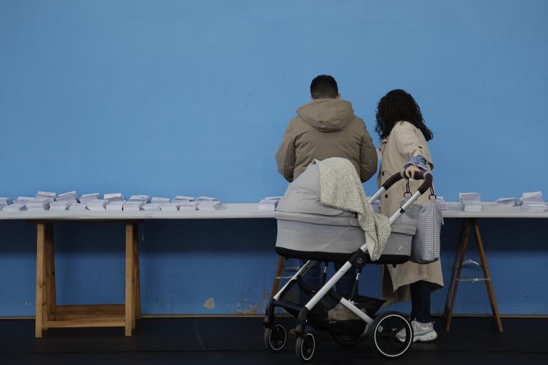 Una pareja con un carrito escoge una papeleta para ejercer su derecho al voto en el colegio Ames a las afueras de Santiago de Compostela