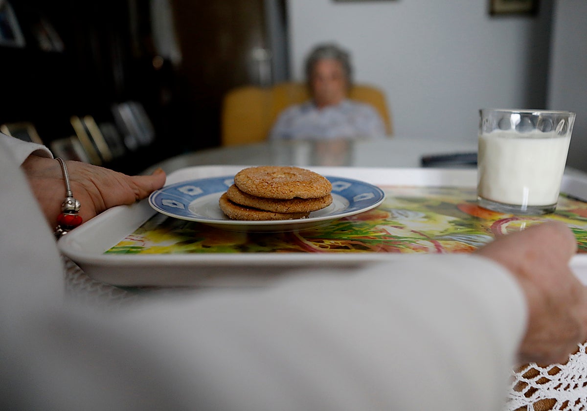 Una asistente lleva una bandeja con leche y pastas a una usuaria de ayuda a domicilio