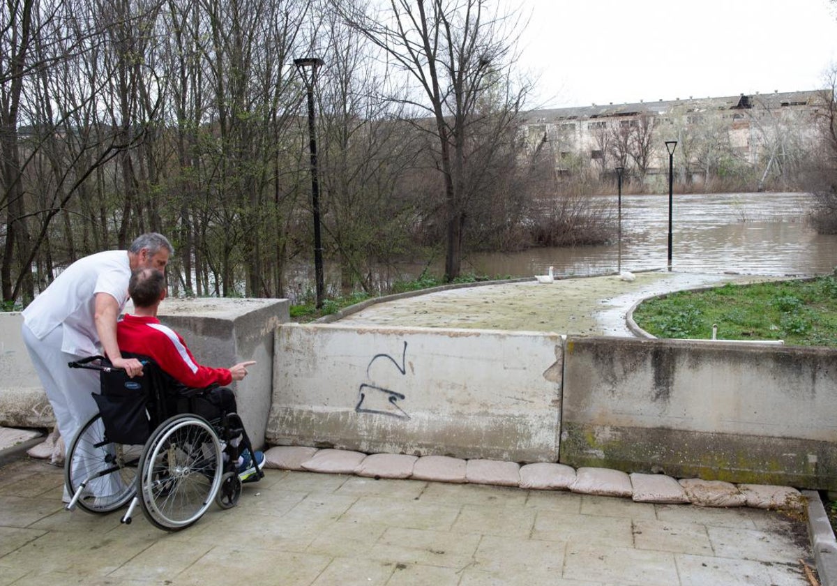 Un paciente de Parapléjicos y su monitor observan el cauce del Tajo, con los paseos del recinto bajo las aguas