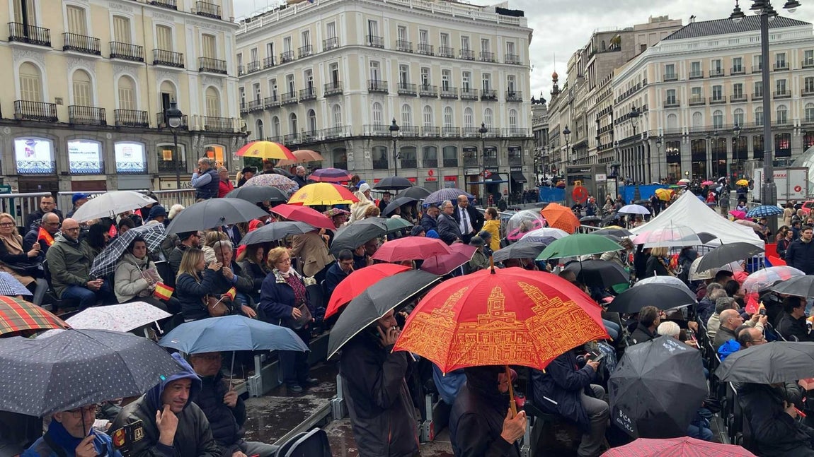 La Puerta del Sol empieza a llenarse a pesar de la lluvia. Se pueden observar diversos paraguas rojos corporativos de la Comunidad de Madrid repartidos entre el público.