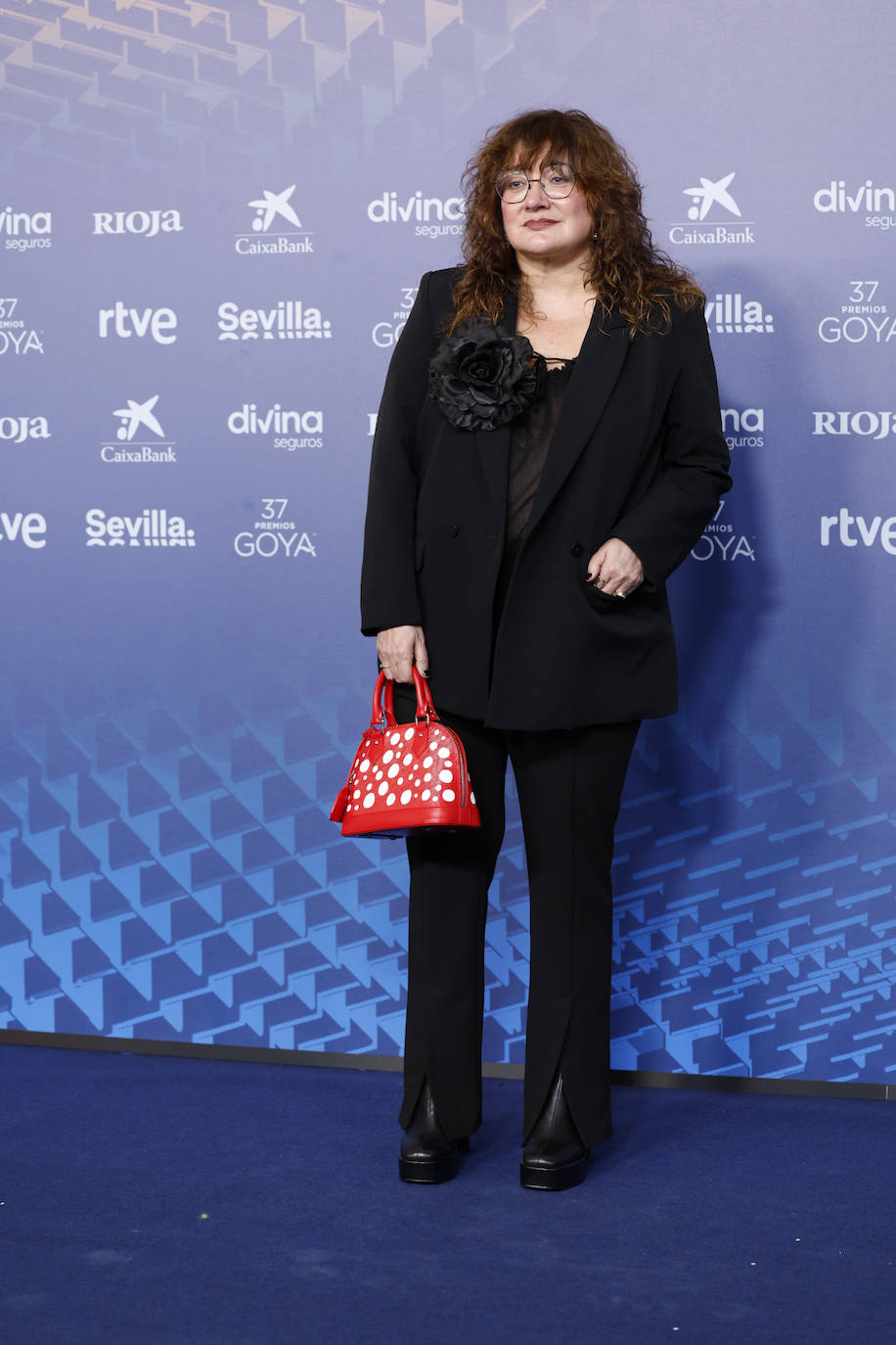 Fiel a su estilo, Isabel Coixet ha elegido un 'total look' en color negro, con pantalón y blazer personalizada con mensaje reivindicativo, pero lo ha combinado con un peculiar bolso rojo con lunares blancos de la colección de Louis Vuitton x Yayoi Kusama. 