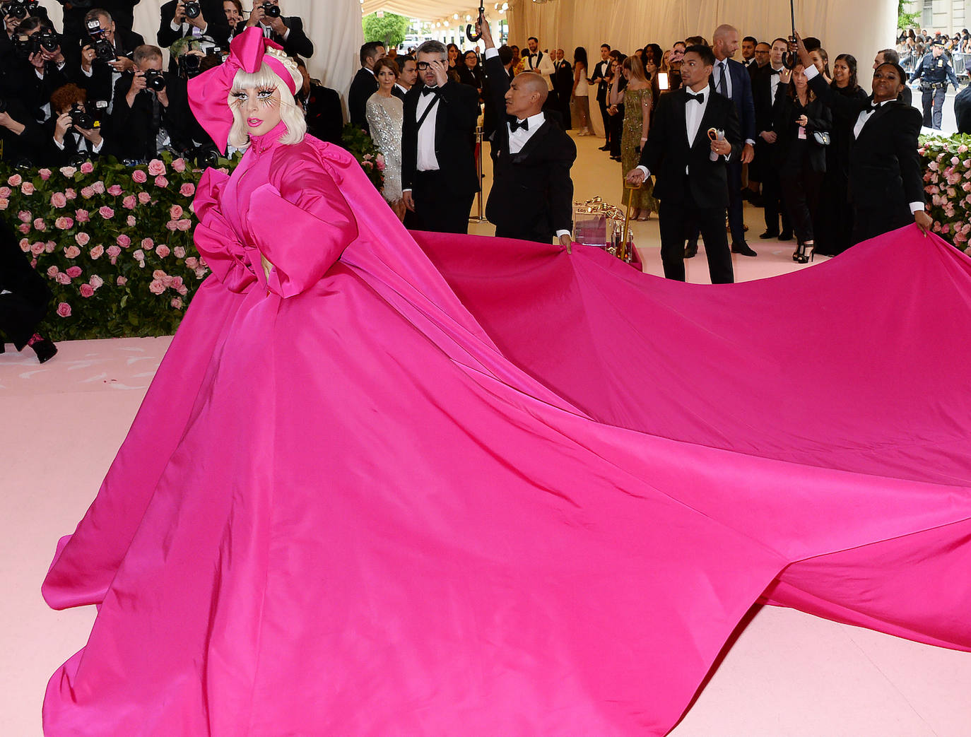 Lady Gaga en la Gala Met 2019 con un look en tono fucsia formado por abrigo-vestido con gran cola y mangas abullonadas del diseñador norteamericano Brandon Maxwell. Lo combinó con maquillaje fantasía con maxi pestañas. 