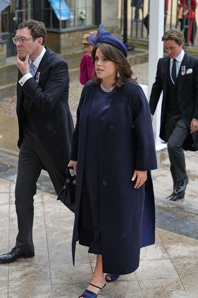 La Princesa Eugenia de York lució un vestido a media pierna de seda y satén azul marino hecho a medida por Fendi by Kim Jones, acabado con un panel lateral de plisse, sobrepuesto con un abrigo de cachemira de doble faz azul marino y azul oscuro de Fendi con un cinturón obi bordado azul marino.   Para completar el look, llevó el bolso Fendi ISeeU Peekaboo de piel azul oscuro con rosas de piel en 3D, tacones Fendi First de piel azul marino y guantes Fendi de seda y satén azul marino bordados con Swarovski.