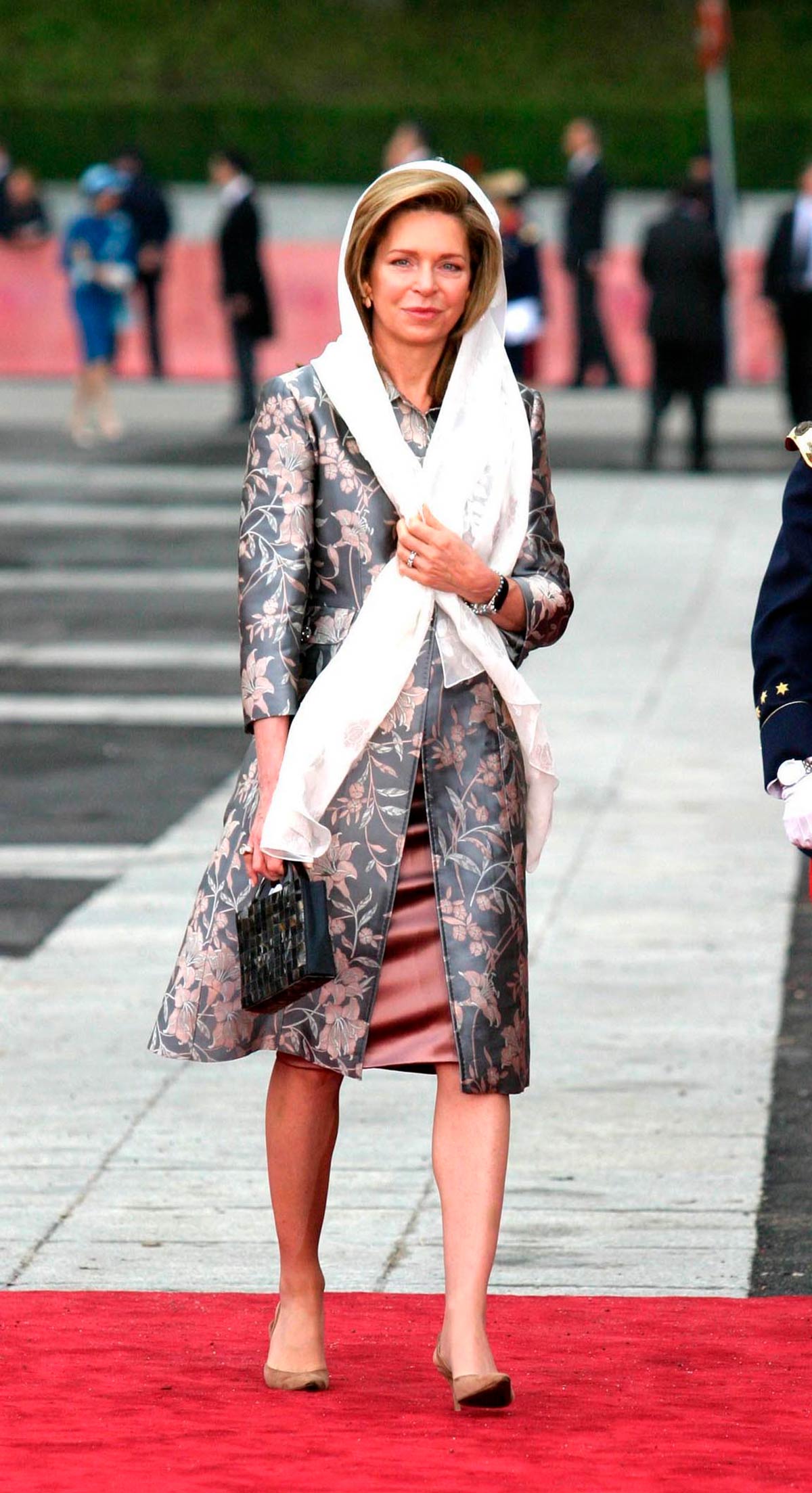 La Reina Noor de Jordania llevó un abrigo gris con flores rosas, un vestido satinado y un pañuelo blanco en el pelo. 