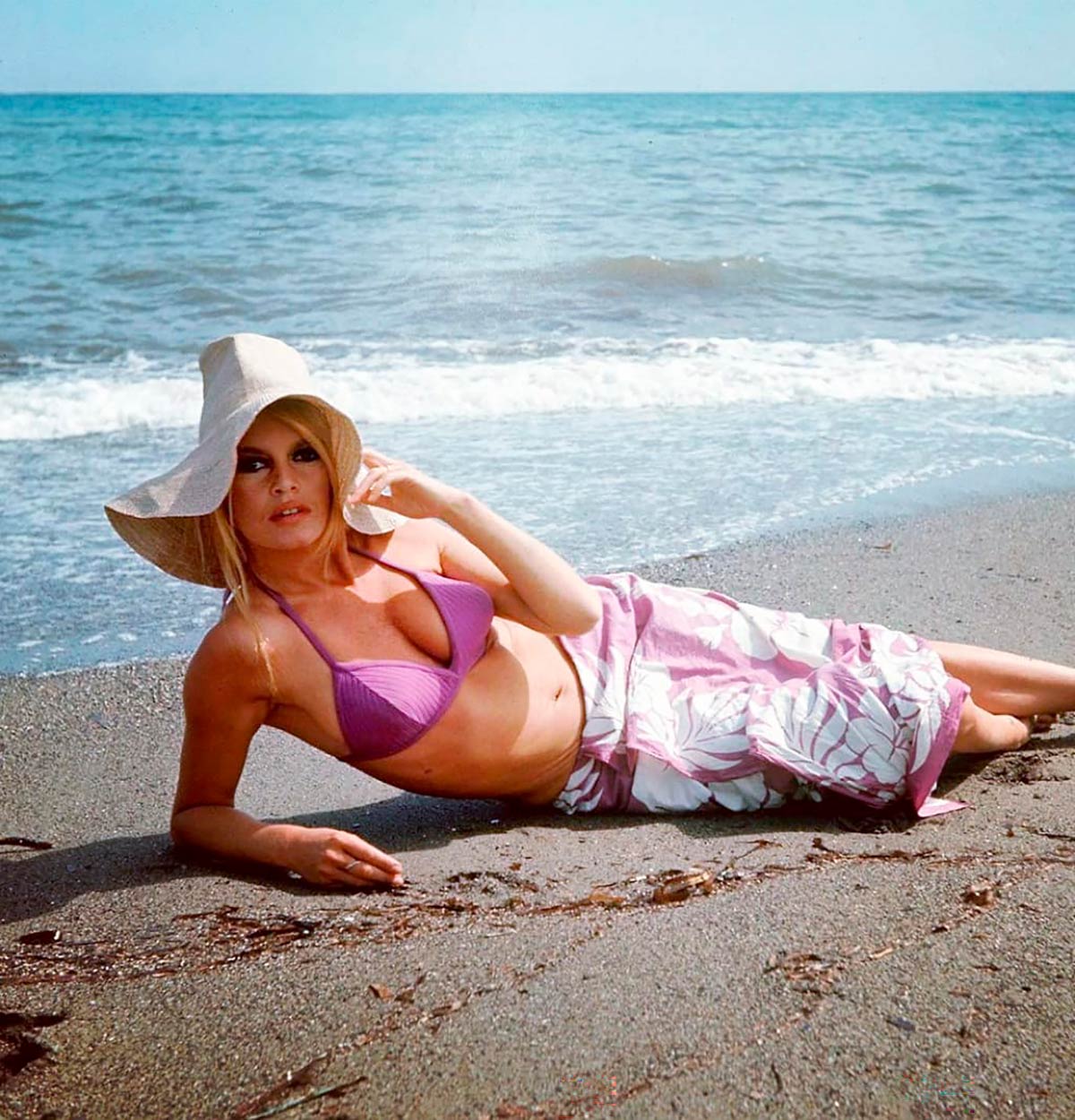 Brigitte Bardot, con un bikini morado triangular, un pareo de flores a juego y una pamela en el pelo. 