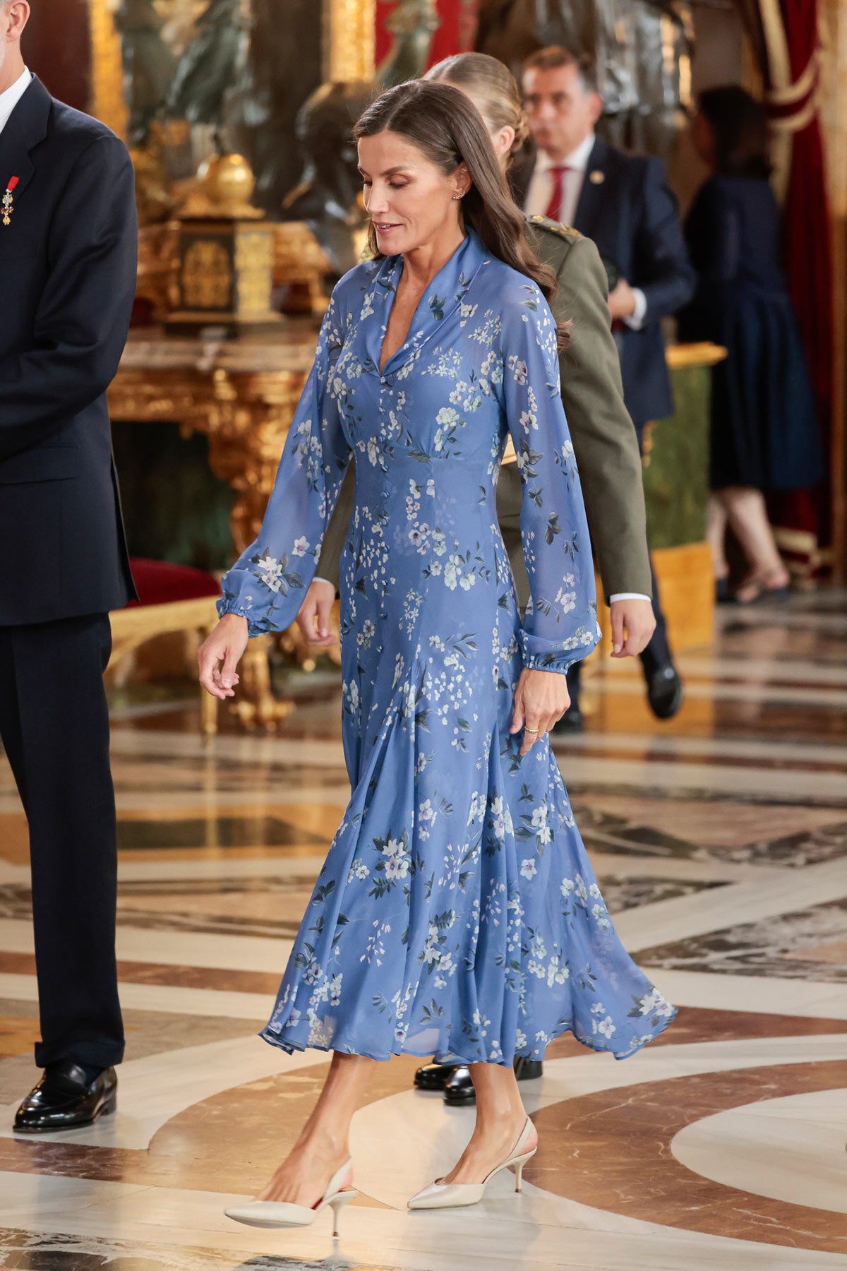 El año pasado, la Reina Letizia estrenó un precioso vestido floral azul del diseñador español Juan Vidal. El modelo, de manga larga y falda de vuelo, presentaba un profundo escote en 'V', frunces en el pecho y una abertura frontal decorada con botones forrados del mismo tejido. Lo remató con unos salones de tacón medio de color beige y joyas discretas. Si bien el Día de la Hispanidad la monarca suele recogerse el pelo, en 2023 optó por dejarlo suelto con ondas ligeras en las puntas. 