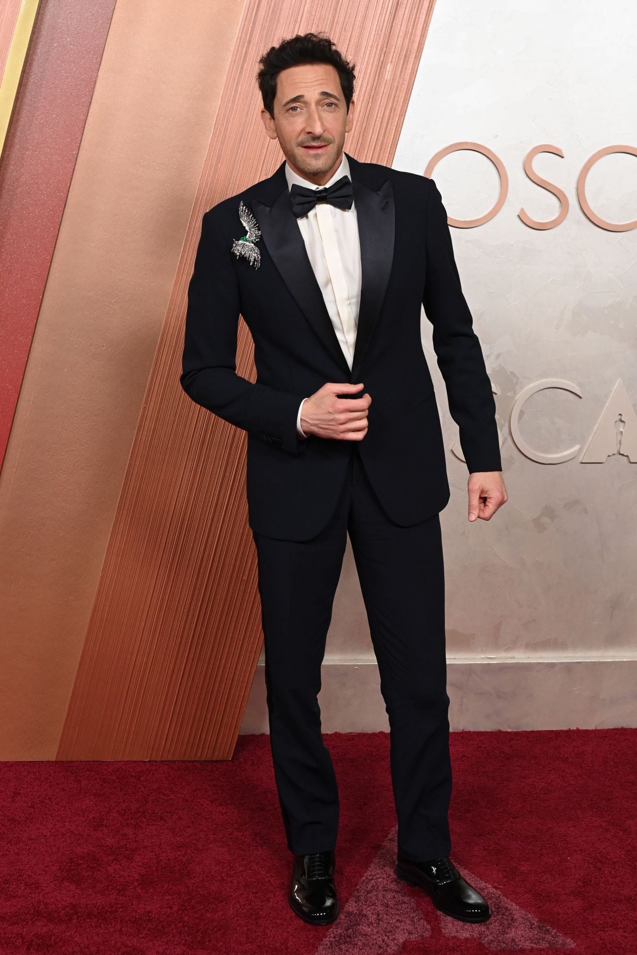 Adrien Brody se ha decantado por el clásico esmoquin negro con pajarita algo más grande de lo habitual y ha puesto el toque de diferenciación con un broche joya de diamantes y esmeralda. 