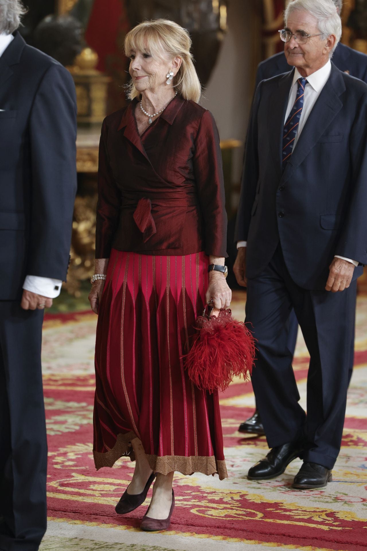 Esperando Aguirre con conjunto de falda y camisa cruzada en tonos burdeos y un llamativo bolso de mano de plumas. 