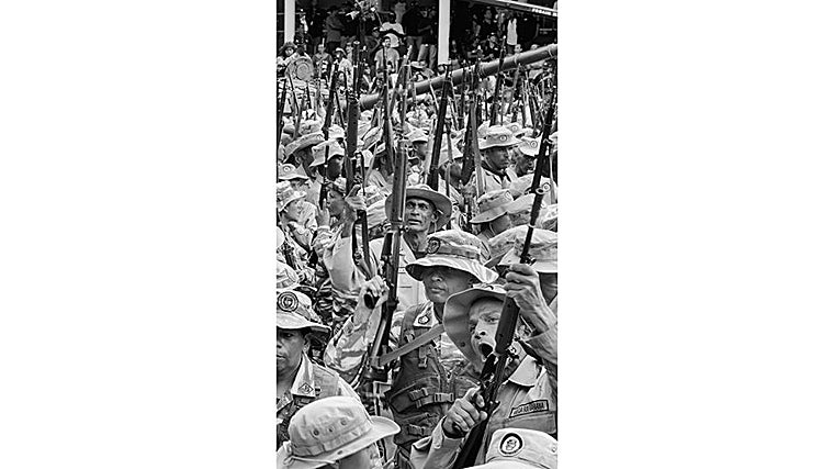 Hombres con el uniforme de la Milicia Bolivariana levantan fusiles en alto durante un acto oficial. Entre rostros curtidos y boinas desajustadas, la disciplina del desfile se mezcla con gestos torpes y cuerpos desordenados