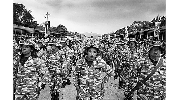 Un grupo de mujeres marcha en formación, la mayoría con apariencia seria, mientras al fondo desfilan cientos de uniformados más. Una de ellas, en el centro, devuelve la mirada directamente a la cámara.