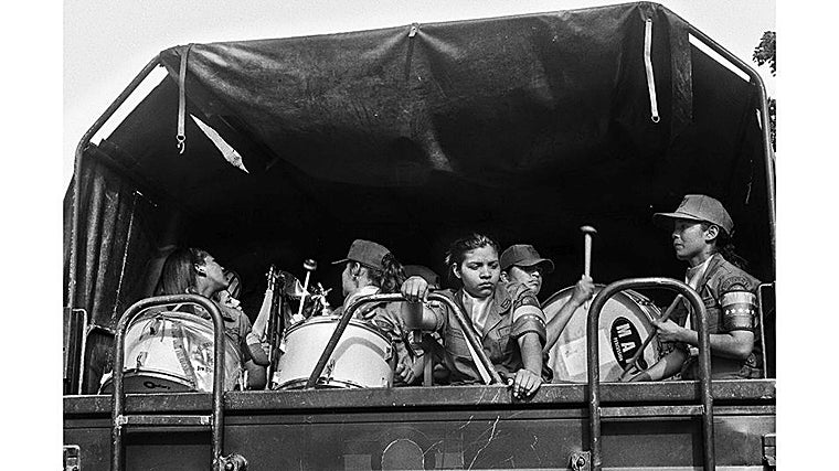 Un grupo de mujeres milicianas viaja en la parte trasera de un camión militar, acompañadas de tambores y redoblantes. Sus uniformes y gorras, perfectamente alineados, contrastan con la expresión cansada y distante de algunas de ellas