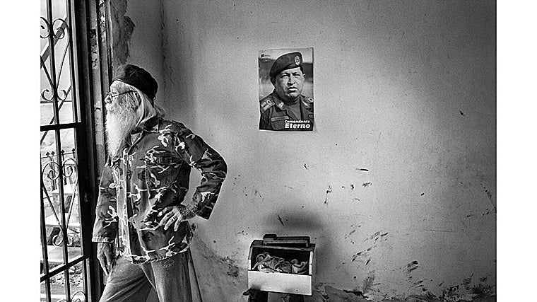 Un anciano con barba blanca y chaqueta de camuflaje observa desde la ventana. En la pared descascarada, un póster de Hugo Chávez lo vigila con la frase «Comandante Eterno»