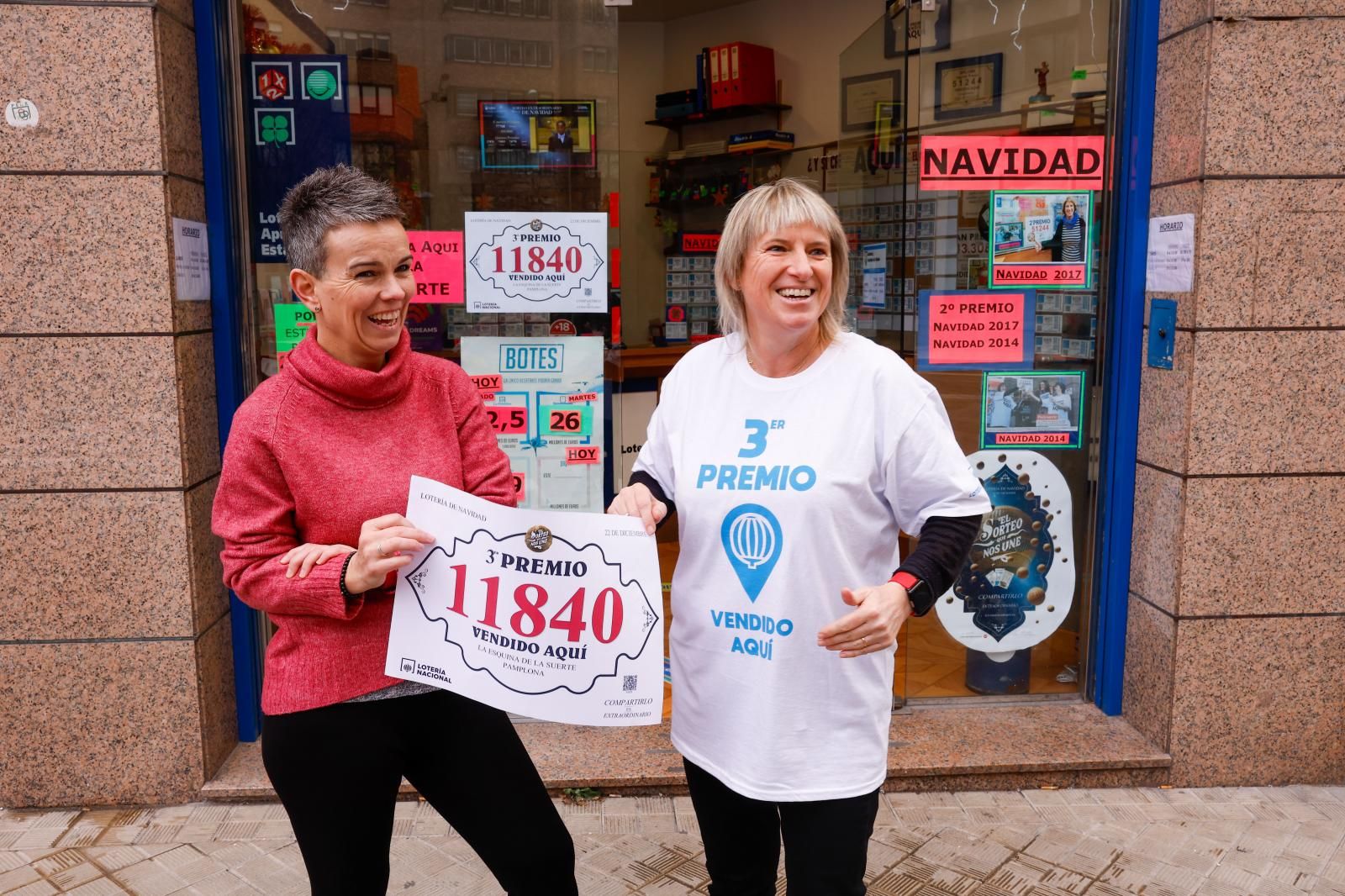 Dos mujeres sostienen un cartel con el número 11840, tercer premio de la lotería, que cae en una administración de Navarra