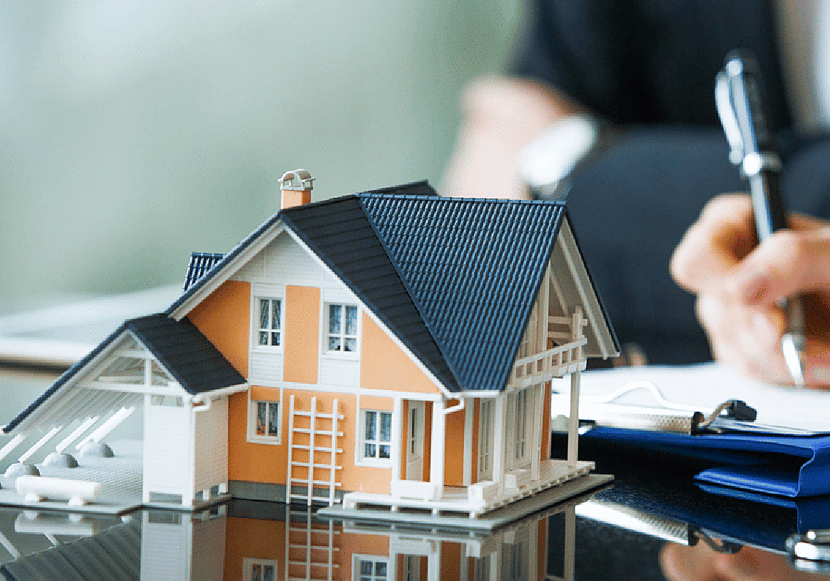 La maqueta de una casa en primer plano con una persona firmando un documento de fondo.