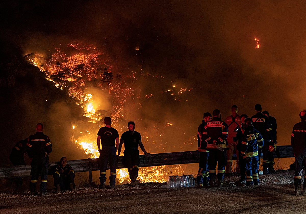 Efectivos del cuerpo de bomberos descansan con el fuego al fondo