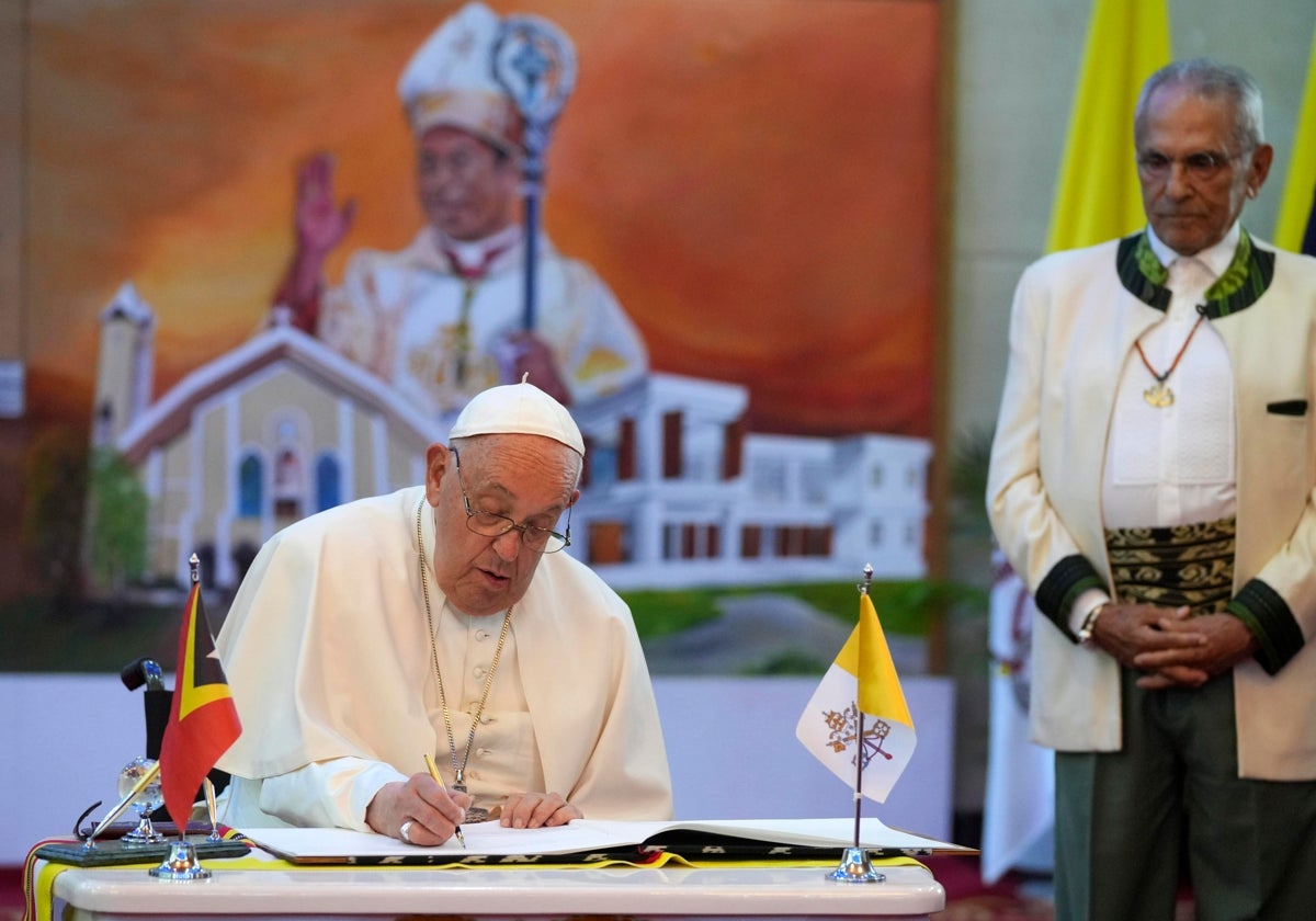 El presidente de Timor Oriental observa mientras el Papa escribe en el libro de visitas durante su ceremonia de bienvenida en el Palacio Presidencial de Dili. En el vídeo, varias personas esperan la llegada del Papa.