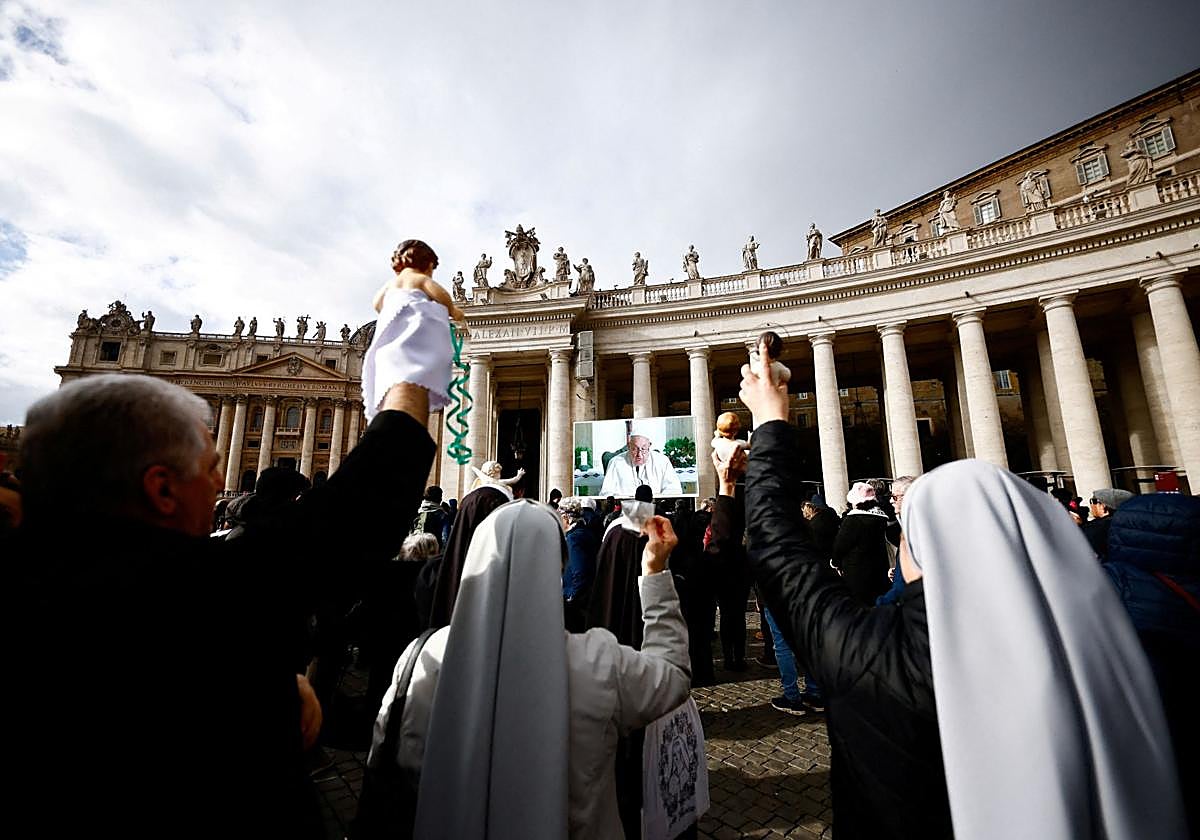 Fieles sostienen figuras del Niño Jesús para recibir la bendición mientras el Papa Francisco aparece en una pantalla dirigiendo la oración del Ángelus desde la capilla de Santa Marta