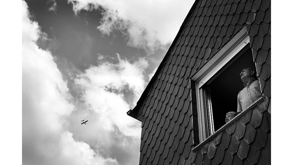 Dos vecinos observan desde la ventana de su casa el paso de un hidroavión. La mirada atenta refleja la preocupación de quienes siguen desde sus hogares la evolución de un incendio que mantiene en vilo a todo el valle. En Fornela, varios pueblos han sido evacuados de manera preventiva y otros permanecen en alerta. La incertidumbre se mezcla con la tensión de no saber si las llamas alcanzarán las viviendas. El paso de los medios aéreos es, para muchos vecinos, la única señal visible de que el esfuerzo por salvar el valle no se detiene.