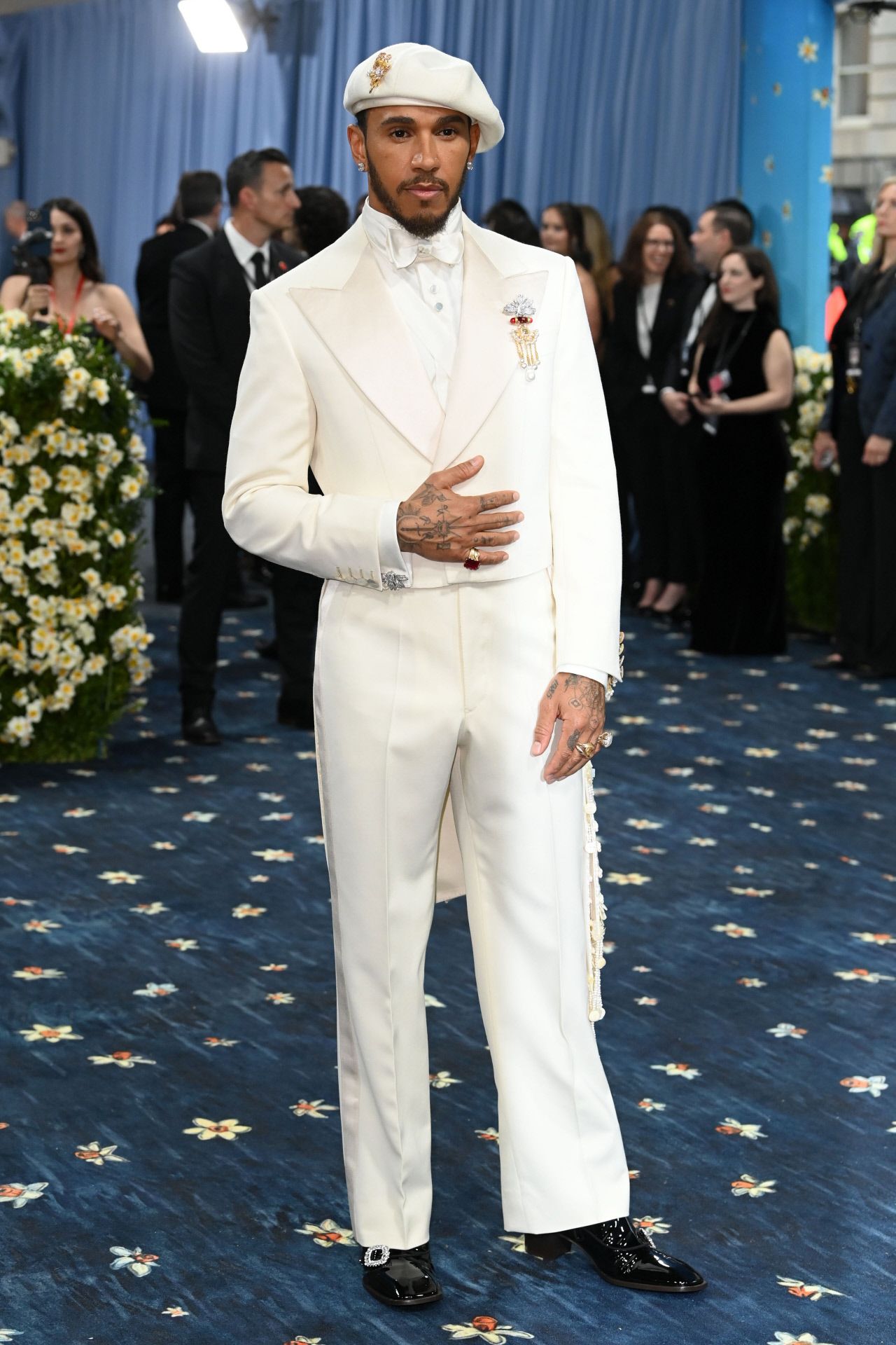 Lewis Hamilton, uno de los anfitriones este año, eligió un traje blanco de la diseñadora inglesa Grace Wales  hecho a medida para la ocasión. Se trata de un conjunto que incluye pantalón recto, americana crop y abrigo. Lo combinó con boina y joyas vintage, incluido un llamativo broche que llevó en la solapa. 