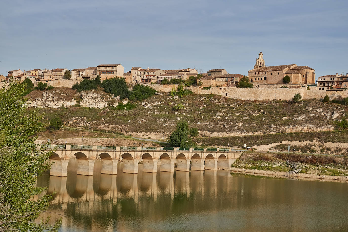 Maderuelo, Segovia.  Un puente moderno ha sustituido al antiguo, cerca de la ermita románica de la Vera Cruz (sus pinturas románicas acabaron en el museo del Prado de Madrid). Al otro lado, rodeando las casas de piedra, se alzan en lo alto del cerro murallas que recuerdan la imagen de un barco. Al pueblo se entra por la almenada puerta de los Dos Arcos, donde nacen las dos calles de la villa. Es tranquilo lugar de casas de adobe y dinteles de piedra, en muchas de las cuales figuran piedras grabadas con extraños signos (flores o círculos), provenientes de la ciudad romana y visigoda que antes hubo. Dos iglesias –una en cada extremo– se reparten las devociones: la de San Miguel y la de Santa María, antigua mezquita según muestran los tres arcos árabes de herradura.