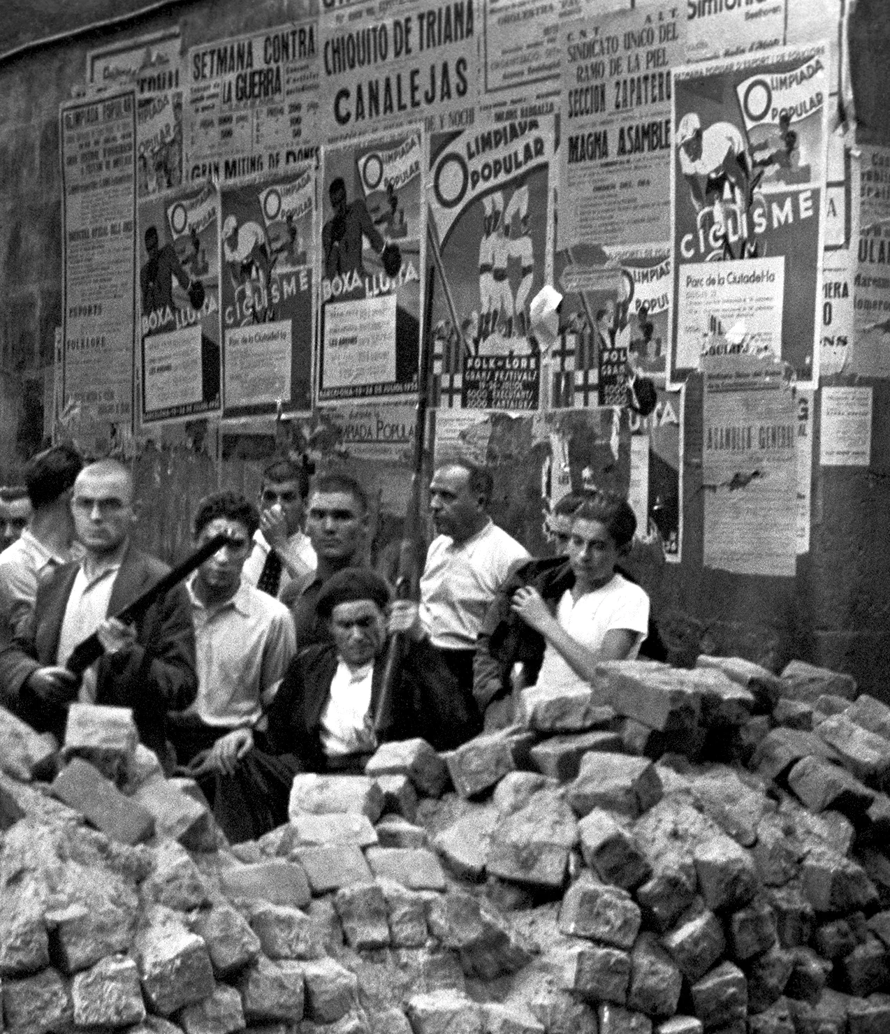 Ciudadanos armados montan guardia junto a una de las primeras barricadas levantadas en las calles de Barcelona, cuyas paredes están todavía adornadas con carteles de la recién cancelada Olimpiada Popular