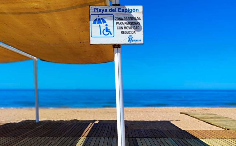 Imagen principal - La playa onubense contará una zona de sombra reservada a personas con discapacidad (arriba). Sobre estas líneas, las ya habituales sillas anfibias y un módulo de aseo.