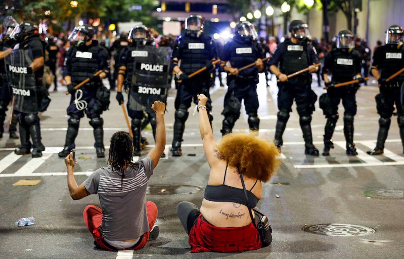 Dos personas se sientan con el puño en altos en frente de agentes que forman un cordón policial