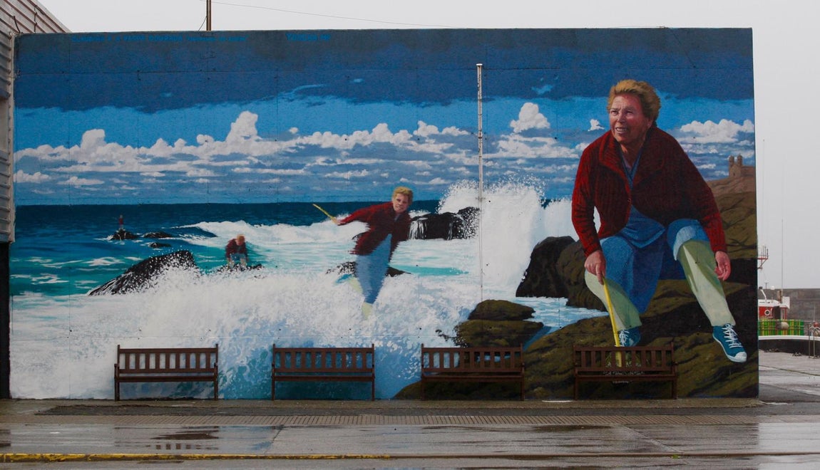 La ninja Claudina. «Costa da Morte, Muxía, rocas y muchas olas, ideal para el percebe. La historía gira en torno a una percebeira, Claudina. Ya su madre había vivido de escapar de la espuma de las olas. Claudina fue al percebe desde los quince hasta los sesenta y cinco, más de una vez casi se la lleva el mar. Pinté a Claudina varias veces en una secuencia saltando de roca en roca como una ninja. La «raspa», que es la herramienta que se usa para rascar en la roca y sacar el percebe, se la pinté de oro. Un excompañero de faena le grita de lejos cuando pasaba por el mural «Claudina te han puesto la raspa de oro», ella, rápida, contesta, «de oro era, que mucho dinero me dio». El mural está en la lonja, ya no se vende pescado pero es el nucleo del puerto donde marineros jubilados, jóvenes mariscadores y niños se mezclan quitandose los sitios de los bancos».