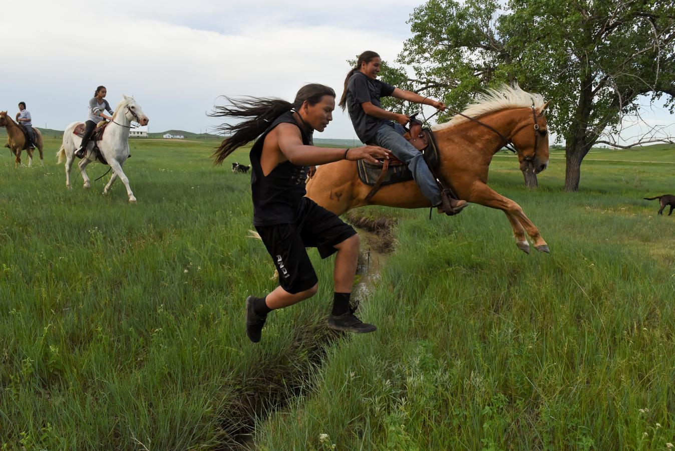 Un hombre salta sobre un pequeño riachuelo a pie mientras su primo salta sobre su caballo en la Reserva del Río Cheyenne en Green Grass, Dakota del Sur, EE. UU., el 31 de mayo de 2018.. 