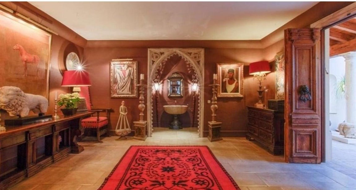 Villa de lujo en Los Monteros. En la estancia de la imagen, de clara influencia árabe, se aprecia un arco apuntado de piedra con ornamentos trabajados al detalle y artesanalmente. Tras ella hay una fuente de piedra bajo un espejo. En la habitación hay varias obras de arte: esculturas de piedra y pinturas. Es la entrada principal y recuerda más a una riad que a una vivienda tradicional española. El tono dominante es el rojo y la arquitectura del espacio es muy personal