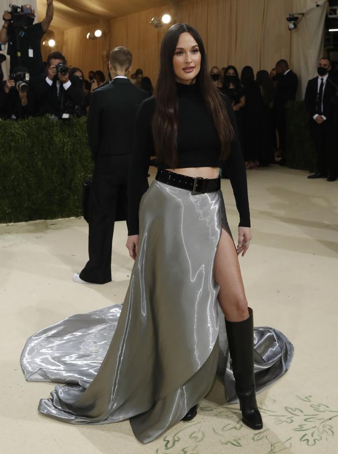 Kacey Musgraves - Alfombra roja de la Gala MET 2021. Como salida de la década de los noventa, eligió una falda metalizada con abertura lateral y un jersey de cuello alzado negro. Cerró el conjunto con unas botas altas negras y un cinturón muy acorde con los que se llevaban en aquella época.