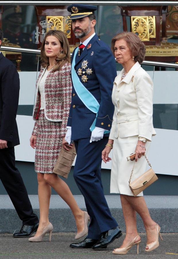 2012 - Los looks de la Reina Letizia en el Día de la Hispanidad. En 2012 también volvió a apostar por el tweed con un traje de falda y americana de tweed en tonos  rojos, verdes y beiges de Felipe Varela que bien podría haber formado parte de una colección de Chanel. Remató su elección con unos salones beige de Magrit y cartera beige de ante firmada por Adolfo Domínguez.