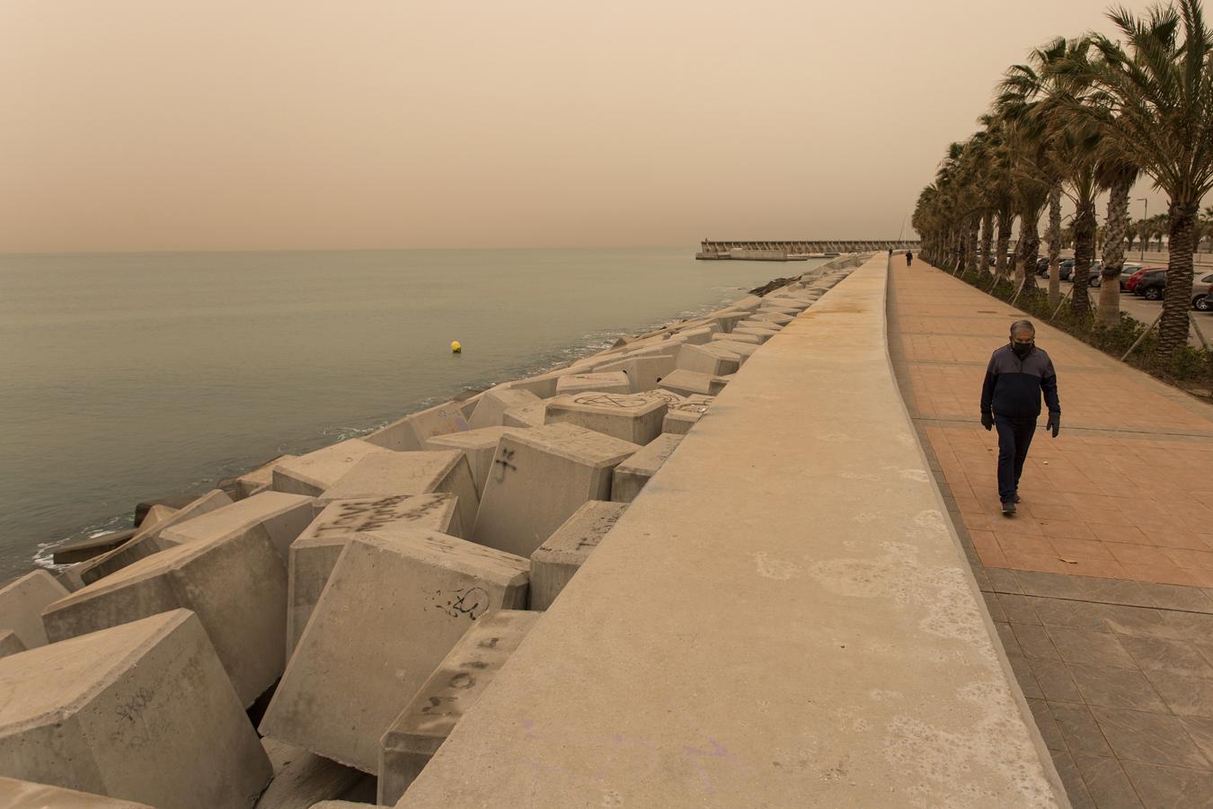 La calima llega a las playas de Málaga. Una persona camina por la zona del Puerto de Málaga que se encuentra cubierta por polvo procedente del desierto del Sáhara que hoy martes ha penetrado en la Península y Baleares, y ha teñido de rojo los cielos de la costa mediterránea