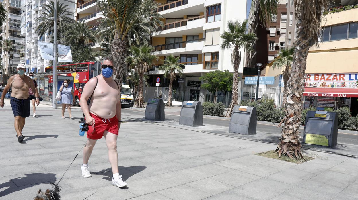 Dos hombres pasean con bañador y mascarilla por el paseo marítimo junto a la Malagueta en Málaga