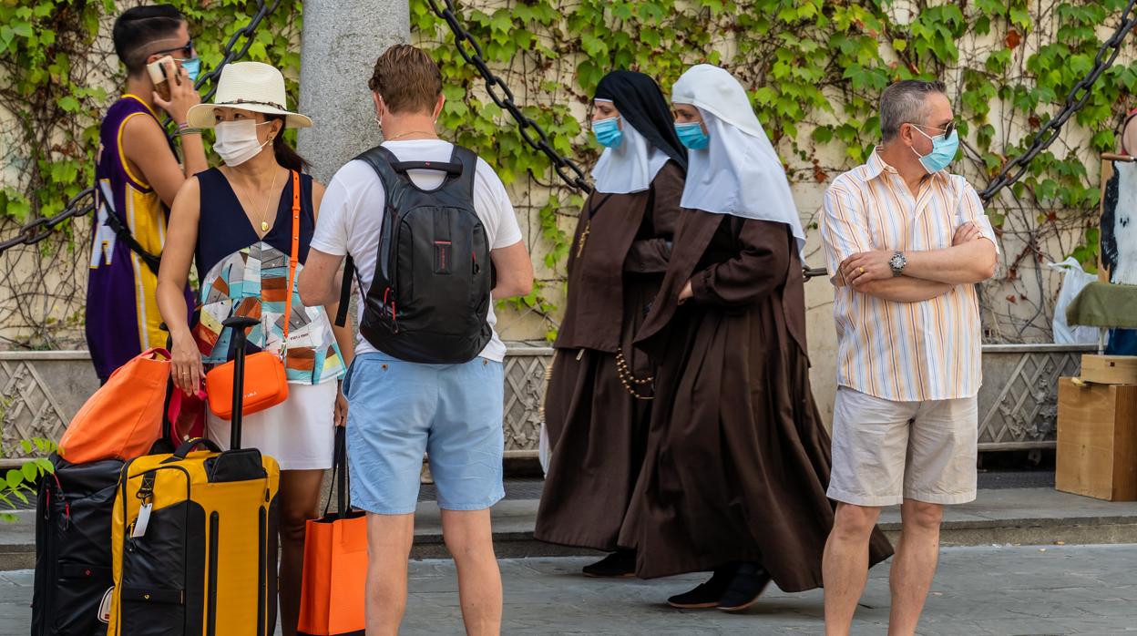 Unos turistas esperan el autobús mientras una pareja de monjas pasan por detrás con mascarillas en Sevilla