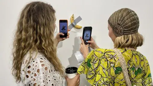 Dos jóvenes fotografían en Basel-Miami «Comedian», el plátano de Cattelan