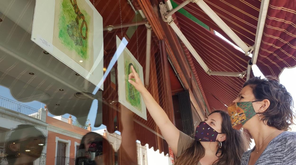 Marta L. Caro y Sheila Criado contemplan las pinturas con las medallas del Rocío de Triana y la Macarena