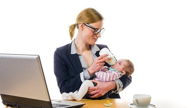 Una mujer da el biberón a su hijo delante de un ordenador de trabajo