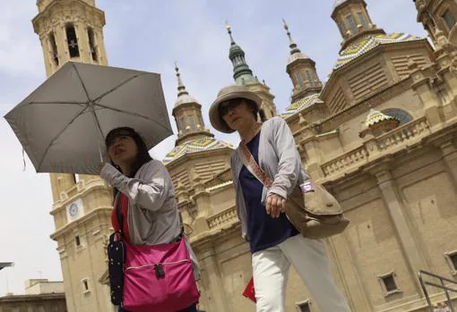 Dos madrugadoras turistas ante la Basílica del Pilar, vestidas por si hacía fresco, acaban protegiéndose del sol porque el calor ya era notable pocas horas después de amanecer