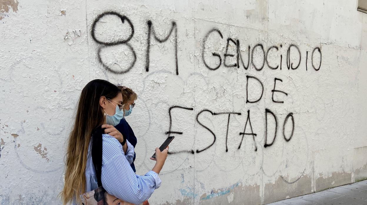 Dos mujeres pasan al lado de una pared en la que hay un graffiti donde se puede leer «8-M genocidio de Estado»