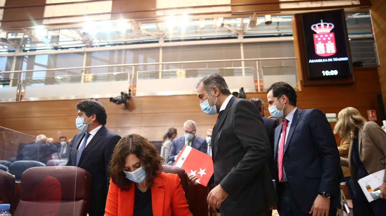 La presidenta Isabel Díaz Ayuso prepara su intervencón en la Asamblea, mientras llegan a sus escaños varios consejeros y el vicepresidente