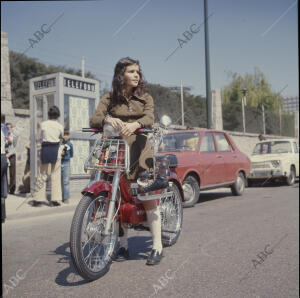 Una joven en una vespino, con unos patines para hielo colgados del hombro, con...