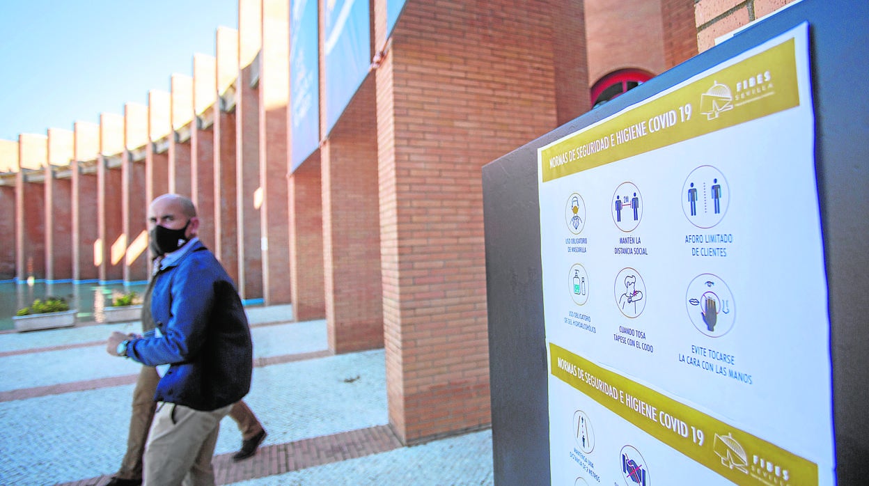 Varias personas salen de Fibes junto a un cartel con medidas sanitarias para evitar contagios