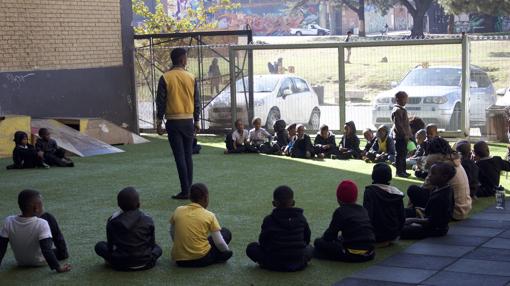 Los alumnos de la escuela Jeppe Park Primary en su recreo, tras la valla otros niños jugando al fútbol en el parque.