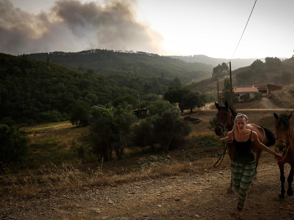 Una mujer tira de dos caballos mientras una columna de humo de alza detrás suyo cerca de la Sierra de Monchique, donde 30 bomberos tuvieron que ser atendidos por los efectos del calor o por inhalaciones de humo.