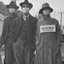 Tres estadounidenses, durante la epidemia de gripe española de 1918, con un cartel advirtiendo de que quien no la lleve ira a prisión